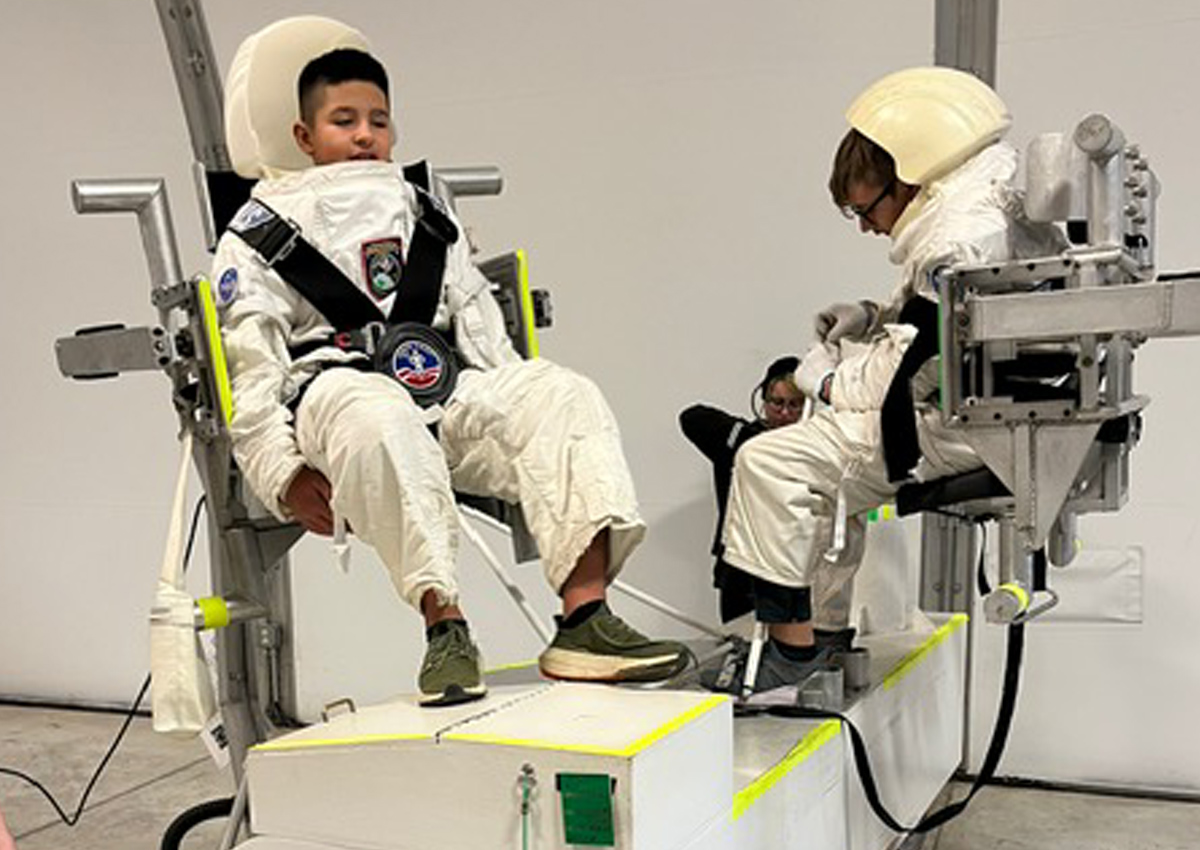 Two adolescents dressed in white, mock, space-suits with open-faced helmets are harnessed into metal zero-gravity chairs attached to the ceiling. One is working on putting something together.