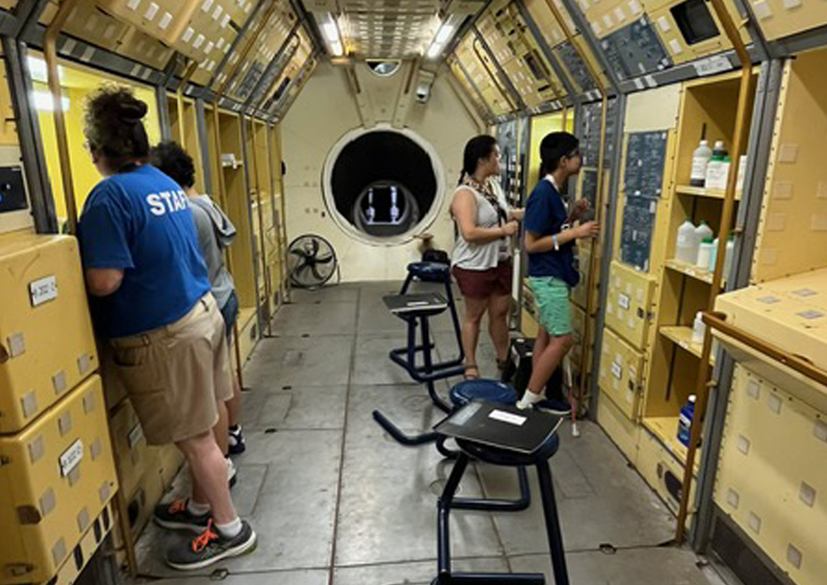A light, mustard yellow, metal with a gray, metal floor. At the far end is a circular opening about three feet in diameter. An adult and adolescent look at a science experiment while two adolescents stand at a work-station conducting an experiment.
