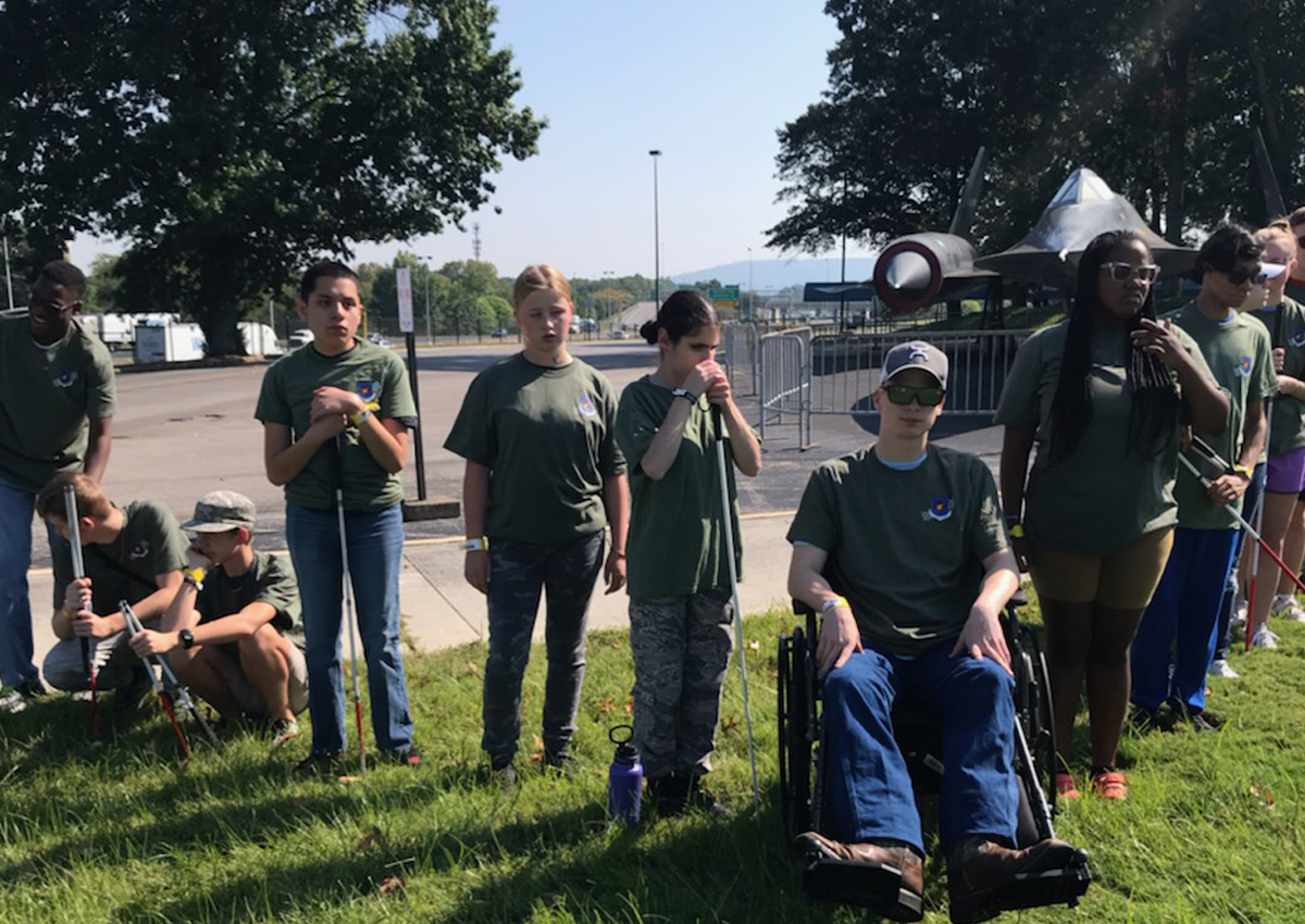 A large group of adolescents in olive green shirts assemble in a v-formation in the grass.