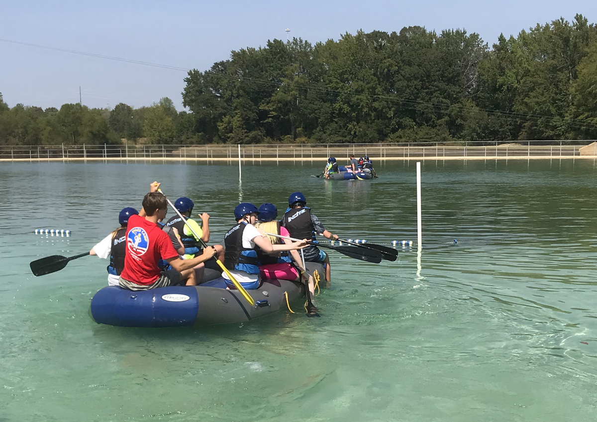 Two large groups paddle inflatable boats toward a goal post in an outdoor pool.