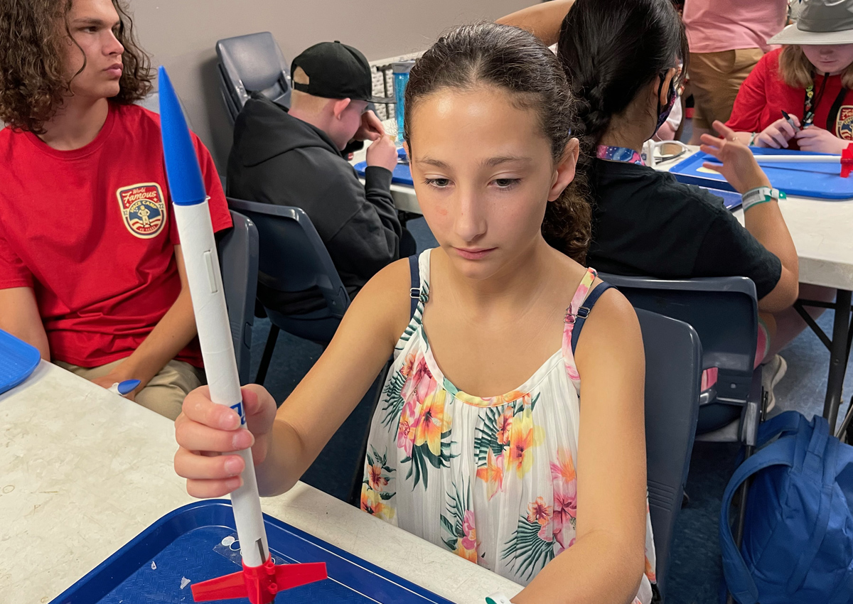 An adolescent holds a model rocket on a blue, plastic tray. Other adolescents build their rockets.