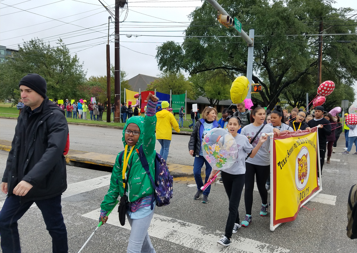 Adults and adolescents hold a TSBVI wildcats banner and helium balloons as they cross a busy street during the White Cane Day march.