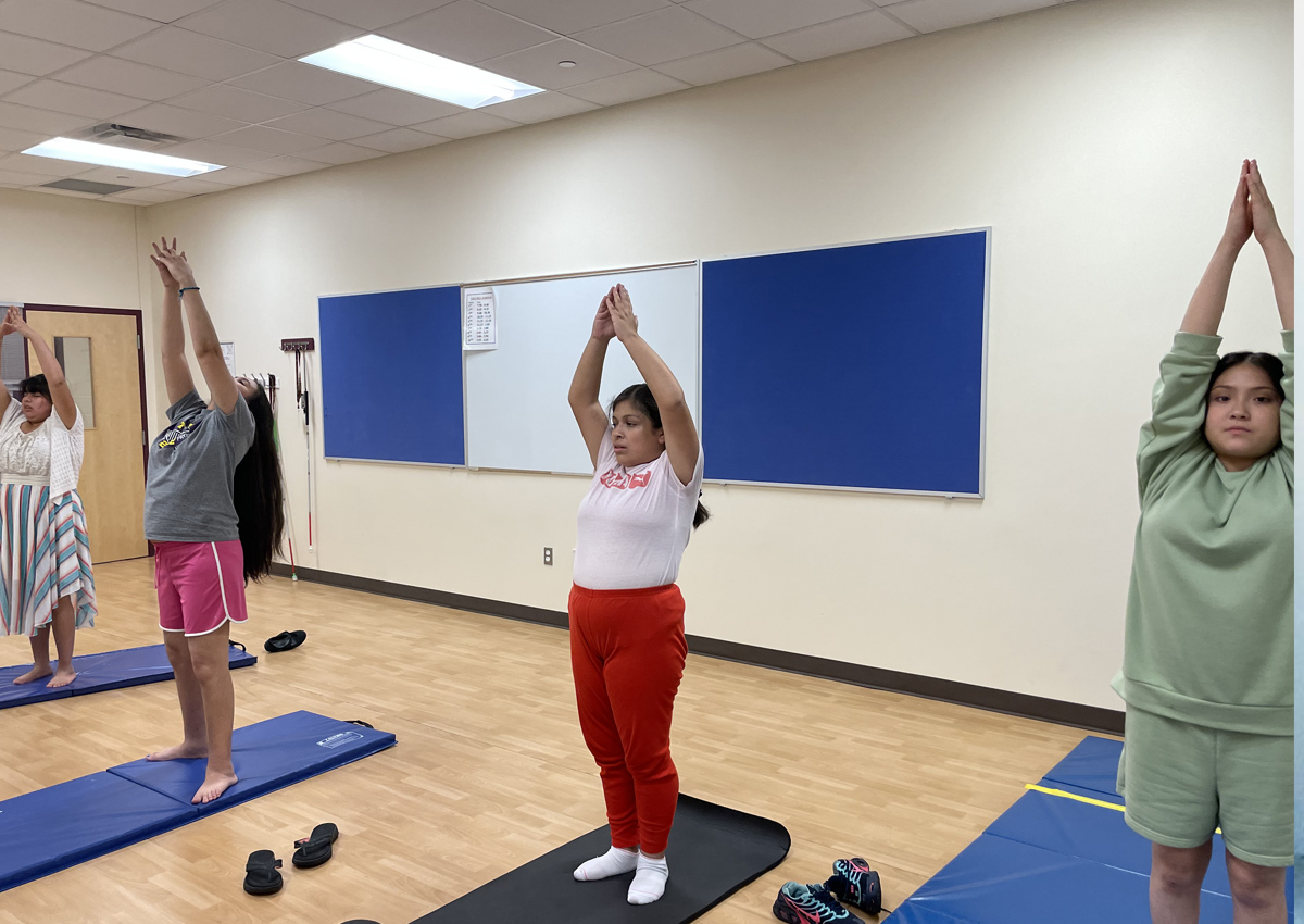 Four adolescents on yoga mats raise their hands above their heads with palms touching.