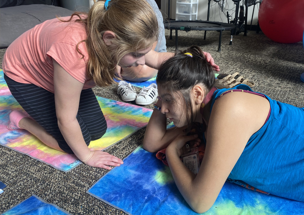 A student kneels on a yoga mat and reaches over to touch the head of her peer who lies on her belly on the yoga mat in a carpeted room.