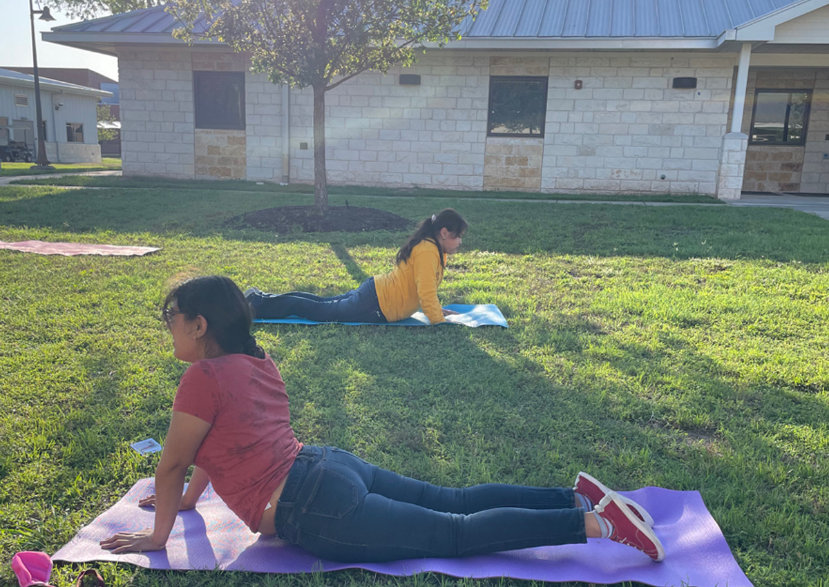 Two adolescents lay on yoga mats socially distanced in a large field in front of a building. They are on their bellies stretching their upper bodies off the mats.