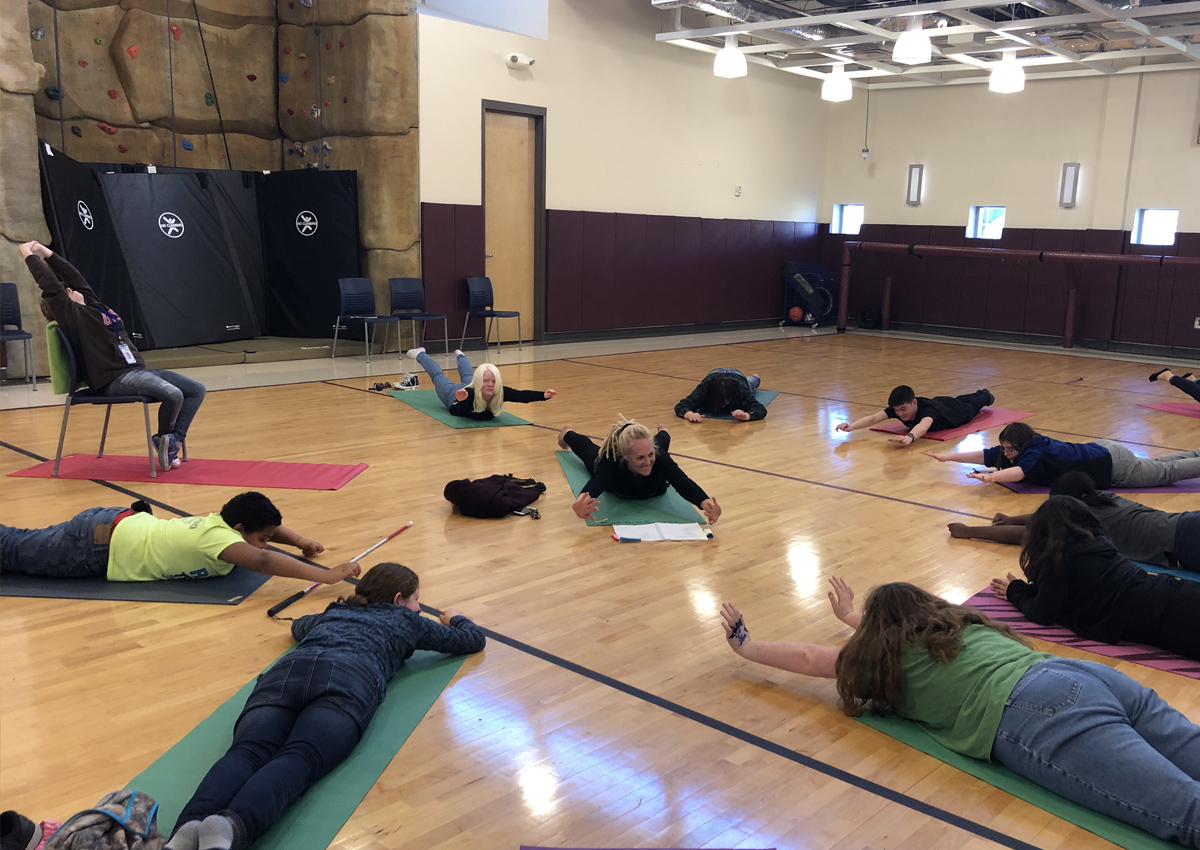 Nine adolescents lie on their bellies on mats in a gym shooting their arms and legs off the mat in varying heights. The mats are arranged in a circle around an adult who demonstrates the pose.