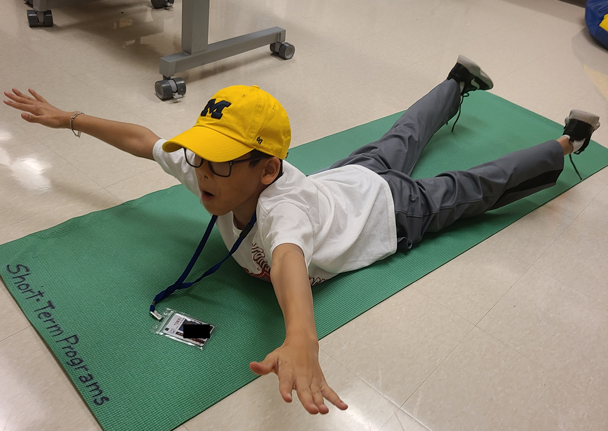 An adolescent on a yoga mat lays on his chest with head and torso up, arms straight out by his sides and legs below the knees off the mat in 