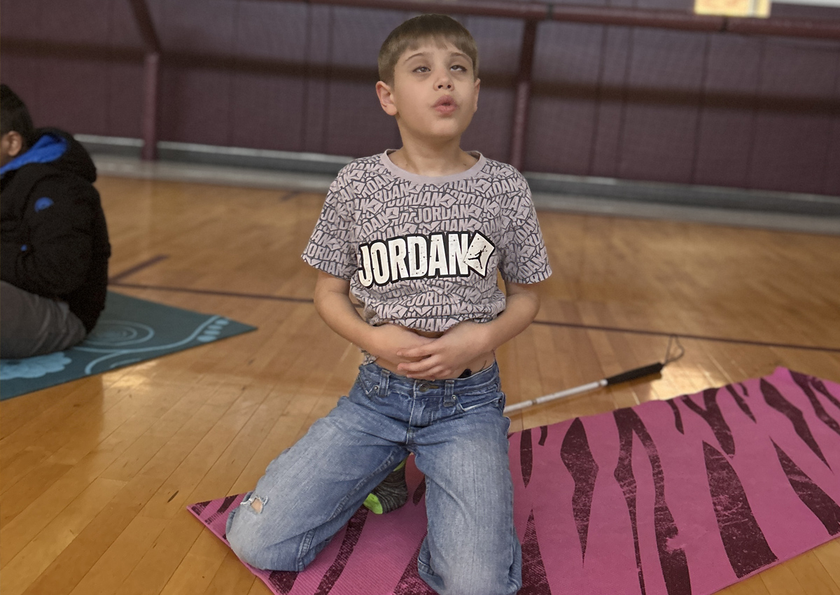 An adolescent sits on his knees on a yoga mat and takes a deep breath while feeling his belly.