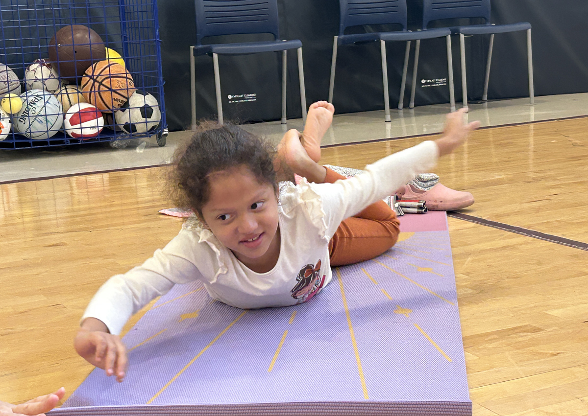 An adolescent lays on her belly on a yoga mat in a gym and stretches out opposing arms and legs.