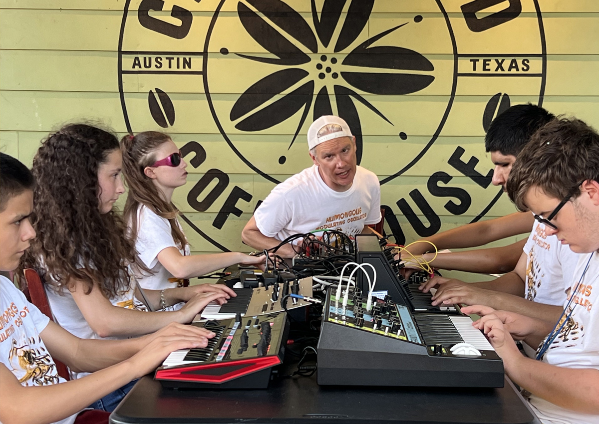 Six adolescents and one adult sit at a table on an outside stage playing synthesizers and sound boards with the words 