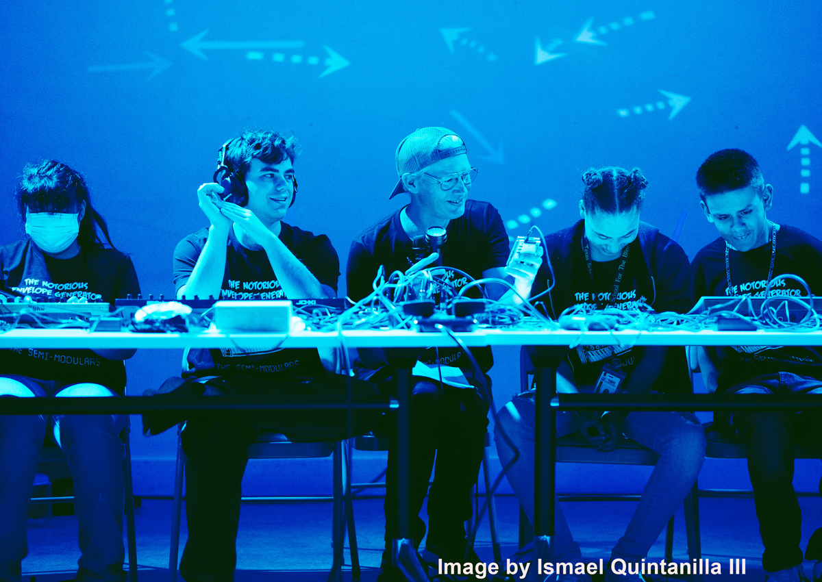 Four adolescents and one adult sit at a table with synthesizers and sound boards inside a dimly lit performance space. Image by Ismael Quintanilla III