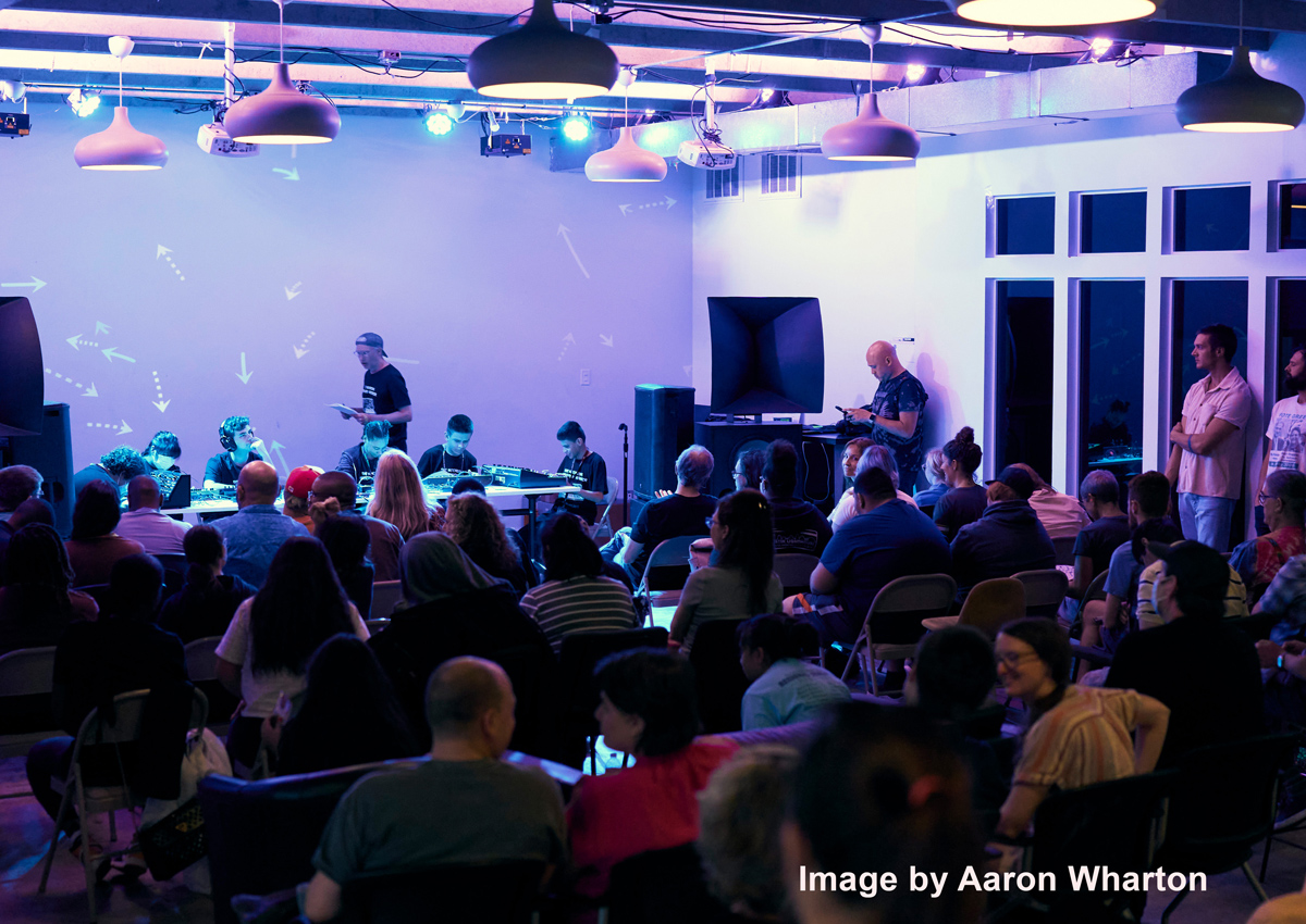SSix students are seated at a long table with analog synthesizers and digital samplers in front of a packed crowd in a low-lit performance space. Image by Aaron Wharton