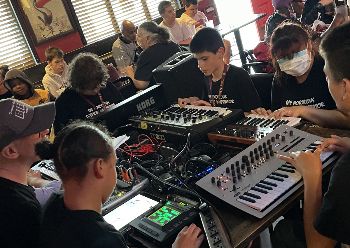 Five adolescents and one adult sit at a table playing synthesizers and sound boards inside a crowded eating venue.