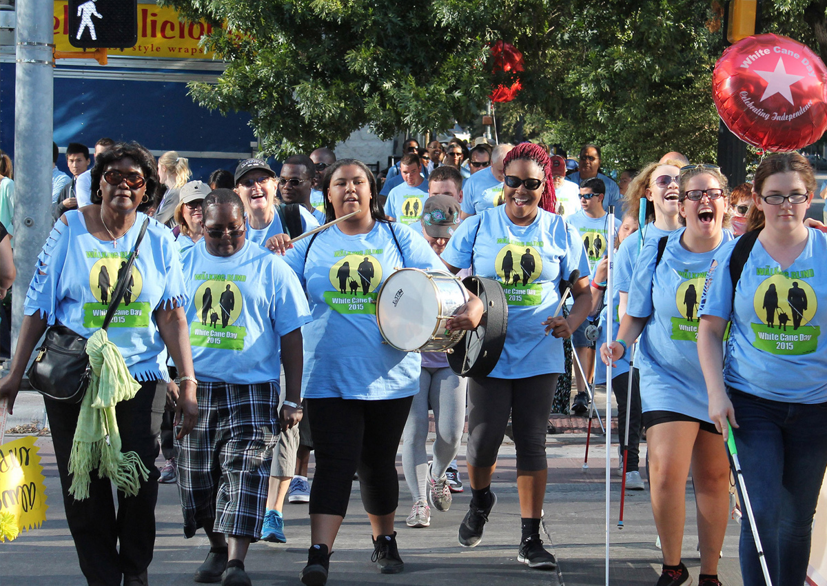 A crowd of adults and adolescents use white canes and wear matching White Cane Day 2015 shirts as they cross a street downtown holding balloons and a snare drum.