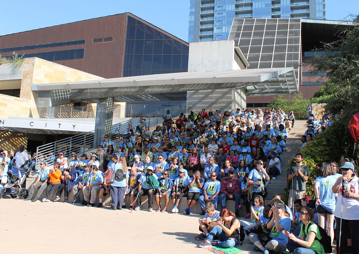 A crowd of people wearing matching White Cane Day 2015 shirts sit on the steps outside City Hall in downtown Austin.