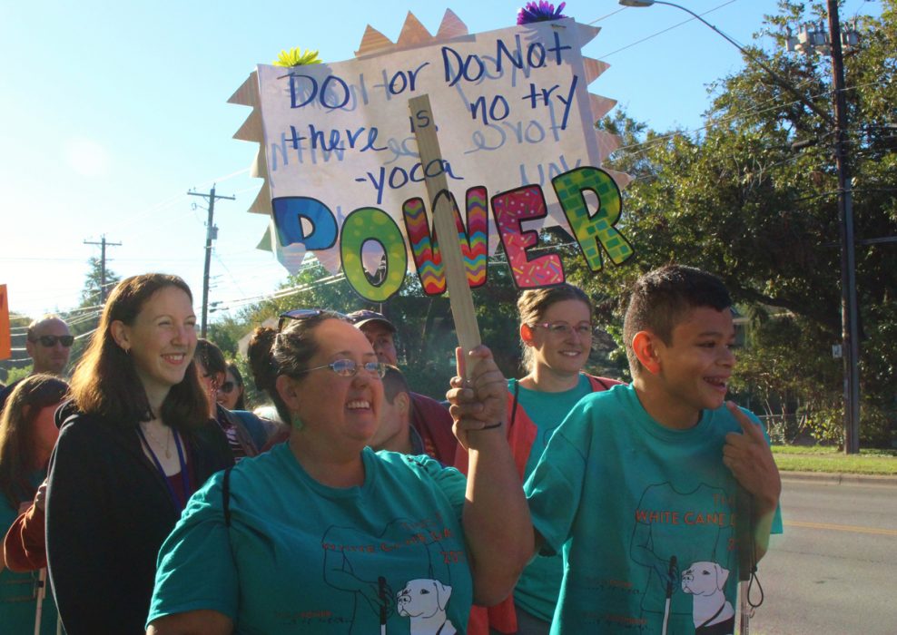 Adults and adolescents wearing matching White Cane Day 2017 shirts march while holding a sign that says “Do or Do Not There is No Try: Yoda Power.”