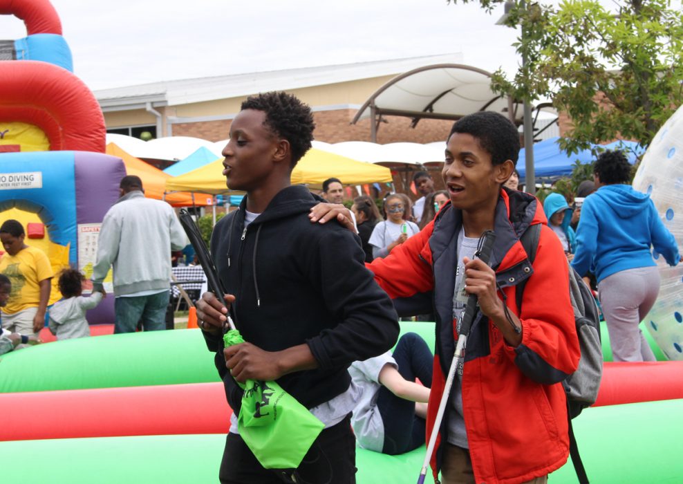 An adolescent holds the shoulder of a peer while walking beside a large bouncy castle with many people outside.