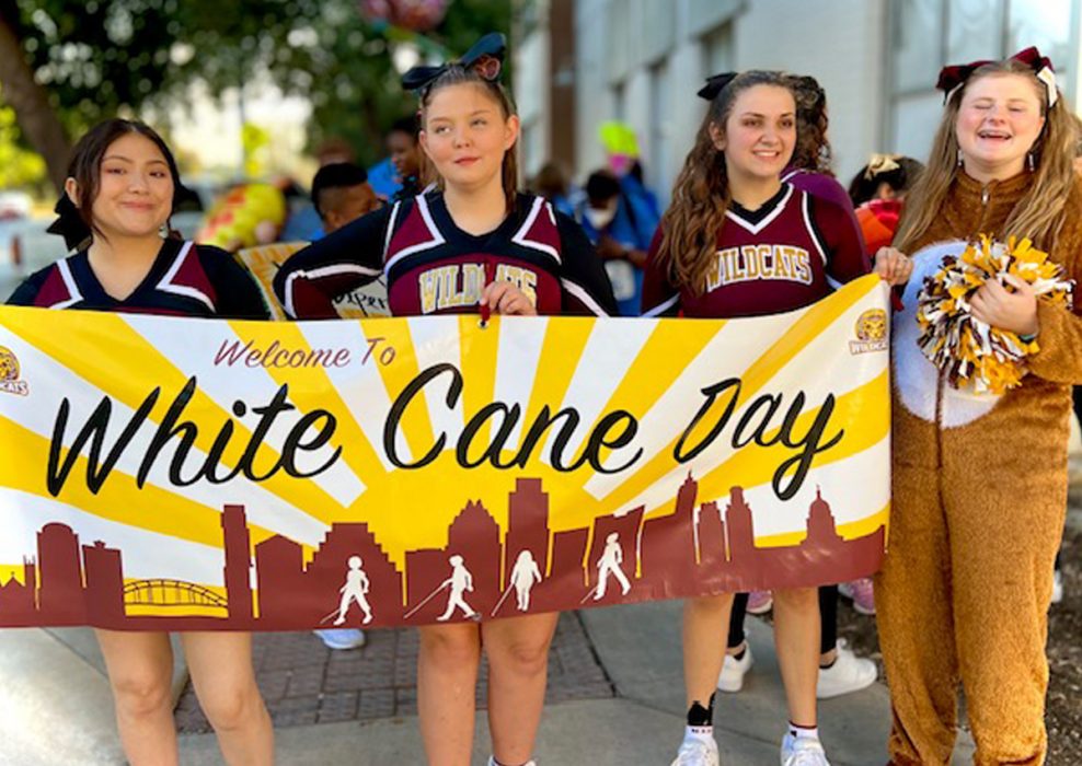 Three adolescents in TSBVI cheerleader suits and a fourth wearing a zip-up furry costume hold a sign “Welcome to White Cane Day” of a cityscape with sun beaming.