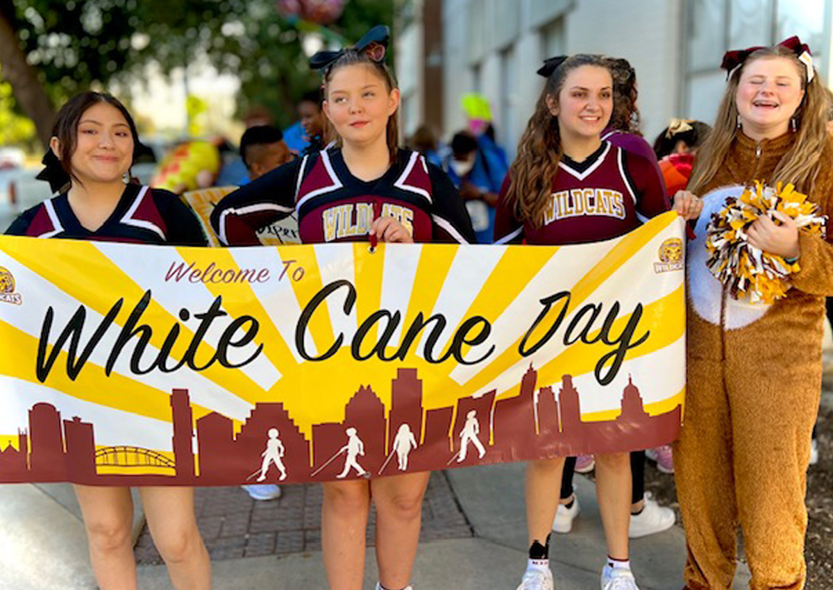 Three adolescents in TSBVI cheerleader suits and a fourth wearing a zip-up furry costume hold a sign “Welcome to White Cane Day” of a cityscape with sun beaming.