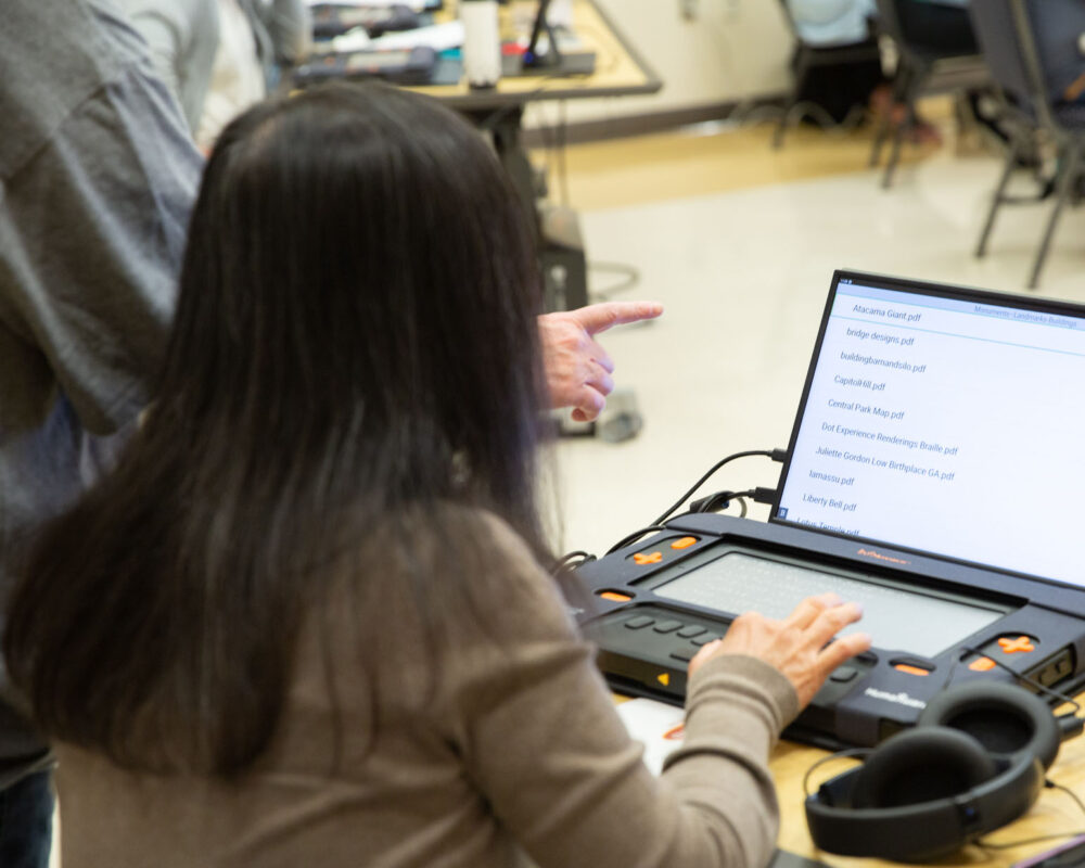 A woman uses a Monarch braille display connected to a monitor.