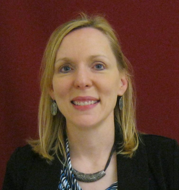 Photo of TSBVI board member Beth Jones standing in front of a red background and wearing a black jacket. She is smiling.