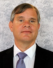 Photo of TSBVI Board member Dan Brown wearing a suit and tie and standing in front of a grey background