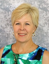 Photo of TSBVI board member Julie Prause. She is smiling and looking at the camera, wearing a blue shirt and standing in front of a grey background.