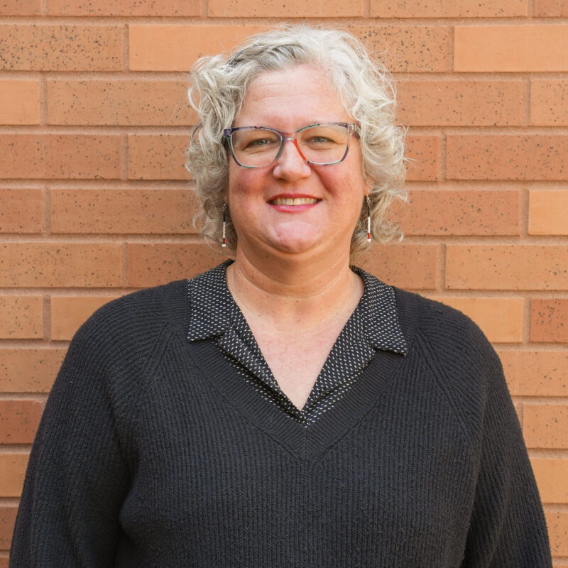 A woman with short curly hair, glasses, and a dark shirt smiles.