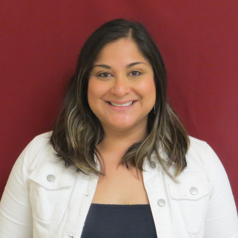 TSBVI Board member Hillary Rodriguez wearing a black shirt and white jacket and standing in front of a maroon background