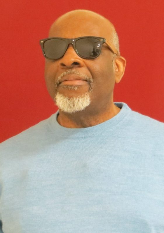 Photo of TSBVI board member Earnest Worthington standing in front of a red backdrop. He is wearing a light blue shirt and sunglasses.