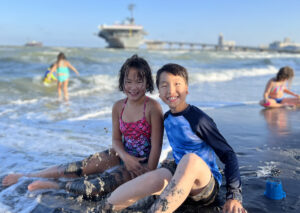 Dos niños en traje de baño sonríen sentados en una playa mientras las olas y la arena les cubren las piernas. A lo lejos se ve un buque de guerra.