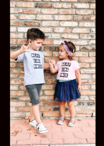 Dos niños con camisetas en las que se lee "Los niños sordos son lo máximo", se apoyan en una pared de ladrillo y se miran. Uno lleva audífono y el otro un implante coclear. Se cogen de la mano y señan "Te quiero".