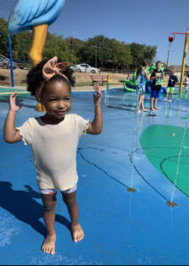 Una niña pequeña con un lazo en el cabello está de pie en medio de un patio de chapoteo con las manos levantadas. Al fondo hay otros niños jugando en el agua.