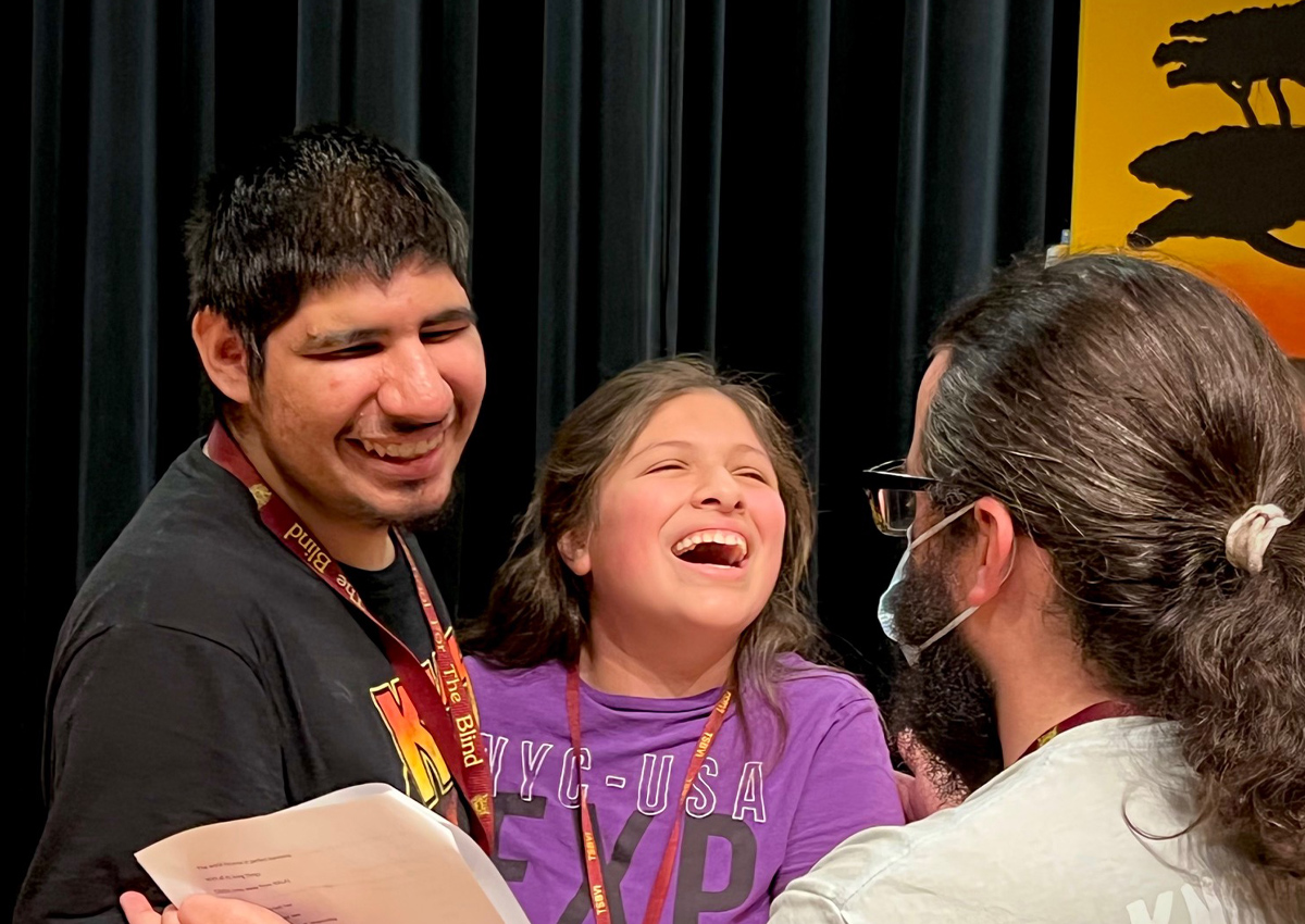 An adult reads from a script in front of two students.