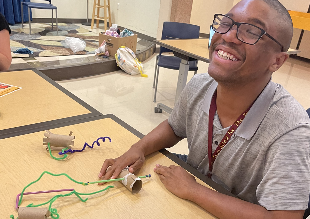 An adolescent seated at a table smiles with pipe cleaners and toilet paper rolls on it.