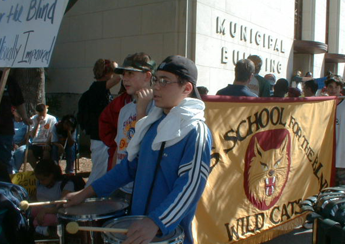 Adolescents outside the old City Hall Municipal Building hold a TSBVI banner and snare drums at the start of a march.