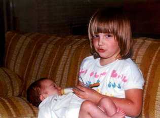 A young girl sitting on couch holding a baby feeding her a bottle