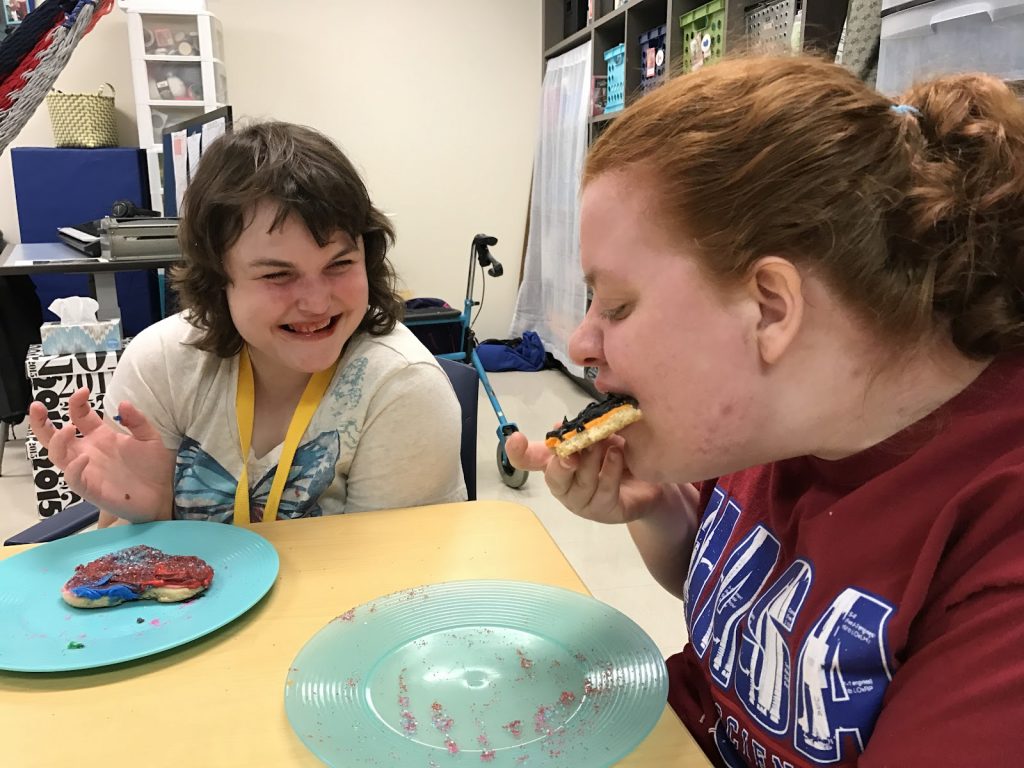 Two smiling students are seated at a classroom table and eating cookies