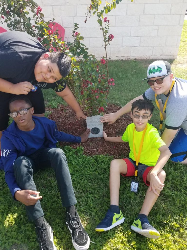 Four students sit around the young crepe myrtle tree, they are all touching the plaque in front of it.
