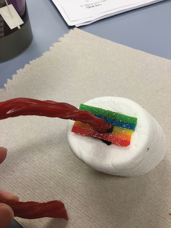 Photo closeup of a giant marshmallow with a rainbow colored sour belt rectangle and a long braided red licorice stick attached to it.