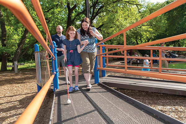 A young girl uses a cane and holds her mother’s hand to explore a park with her parents.