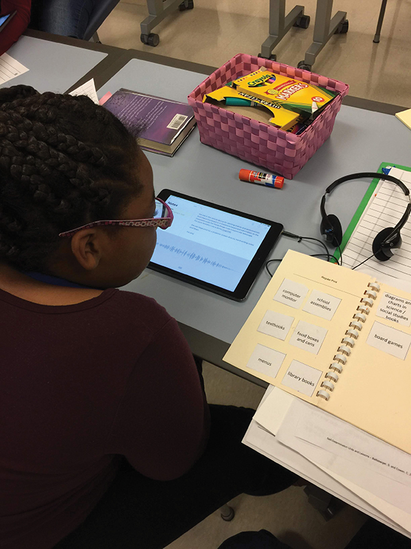 Picture of a student sitting at a desk with an iPad and school supplies. Her Current Access Book is opened to the “Regular Print” page with tasks such as board games, library books, menus, and diagrams.