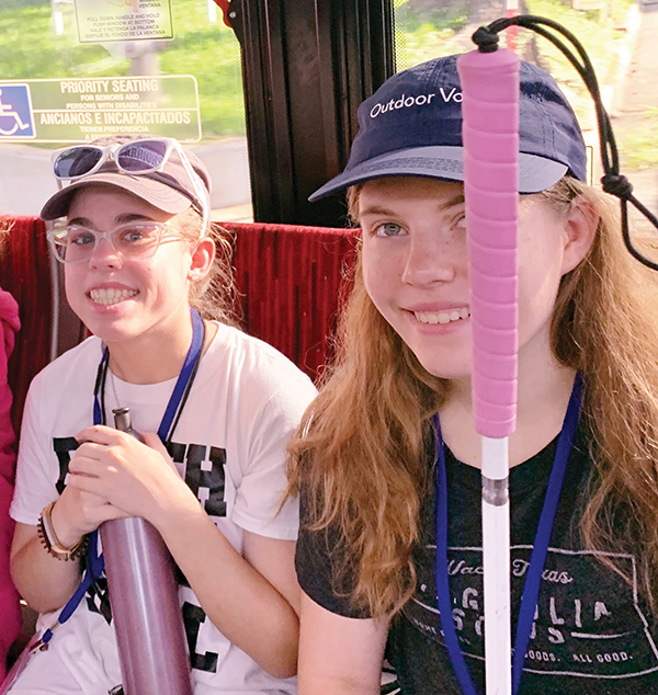 Two girls ride the city bus. One student is wearing glasses and carrying a water bottle. The other student is wearing a hat and has her long white cane with a pink grip.