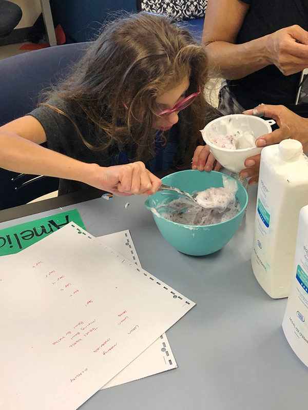 A girl with long hair and pink glasses sits at a table scooping lotion into a bowl. To her right is the braille recipe she’s using to make soap.