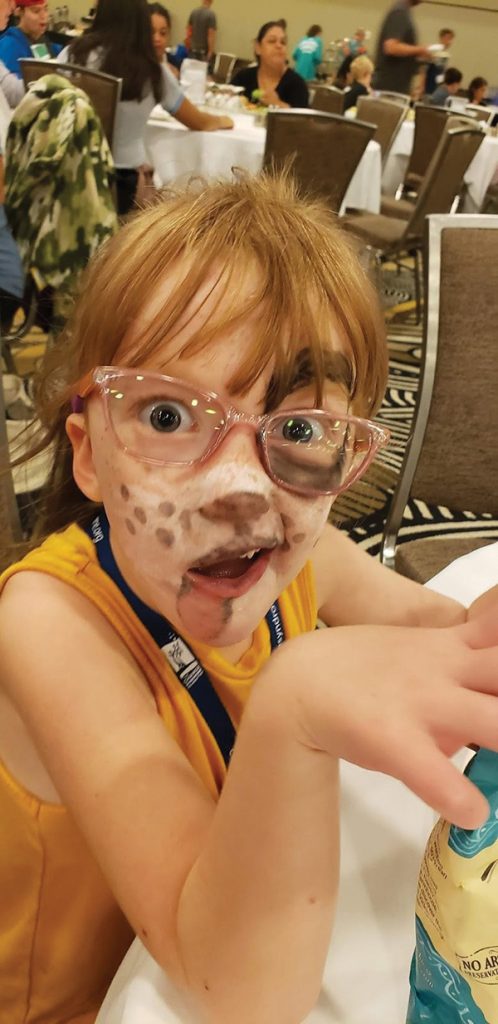 A little girl with CHARGE syndrome smiles for the camera. Her face is painted like a puppy.