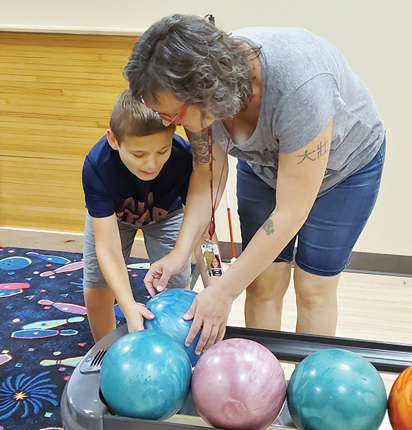 A boy and his teacher lean over the bowling ball return to pick up a blue bowling ball.