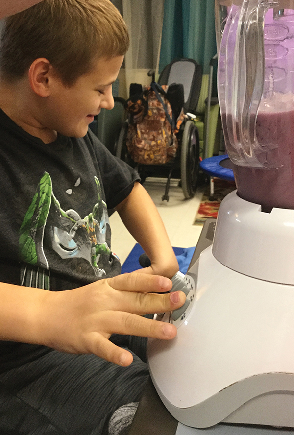 A boy sits in front of a blender containing a partially blended smoothie. His fingers are on the buttons of the blender, and he is smiling.