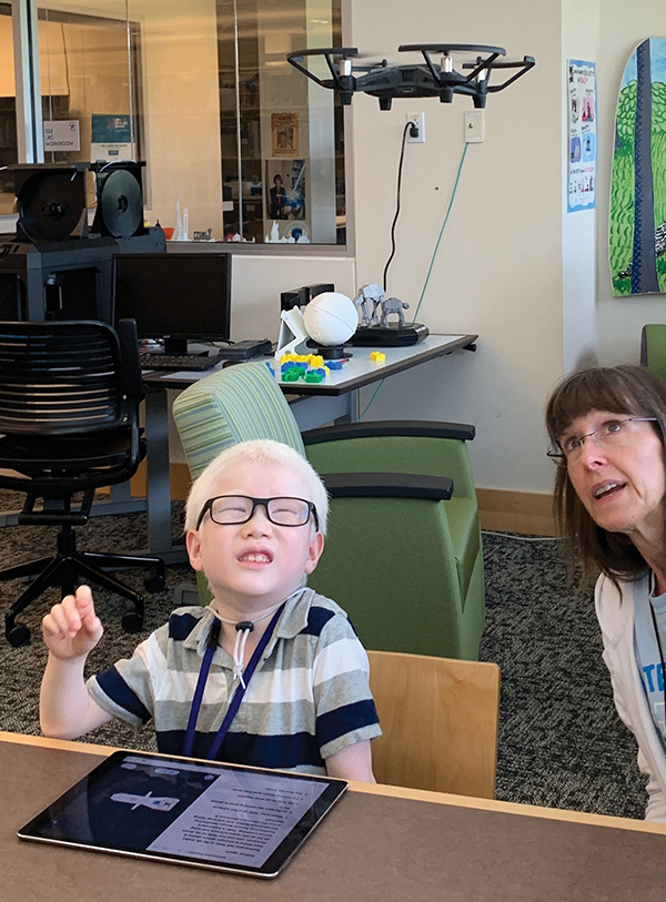 A young boy and a staff member sit at a table with an iPad in front of him. He is looking up at a black drone hovering above him.