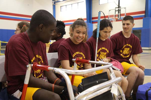 Players sitting on a bench in the TSBVI gym, conversing while taking a break between matches. One student uses a walker and another has a cane.