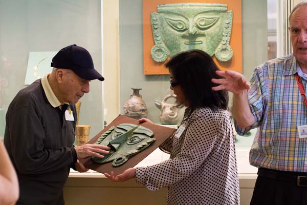A docent holds a three-dimensional replica of a piece of art for a patron to explore with his hands. The original, a large green mask, is pictured in the background, preserved behind glass.